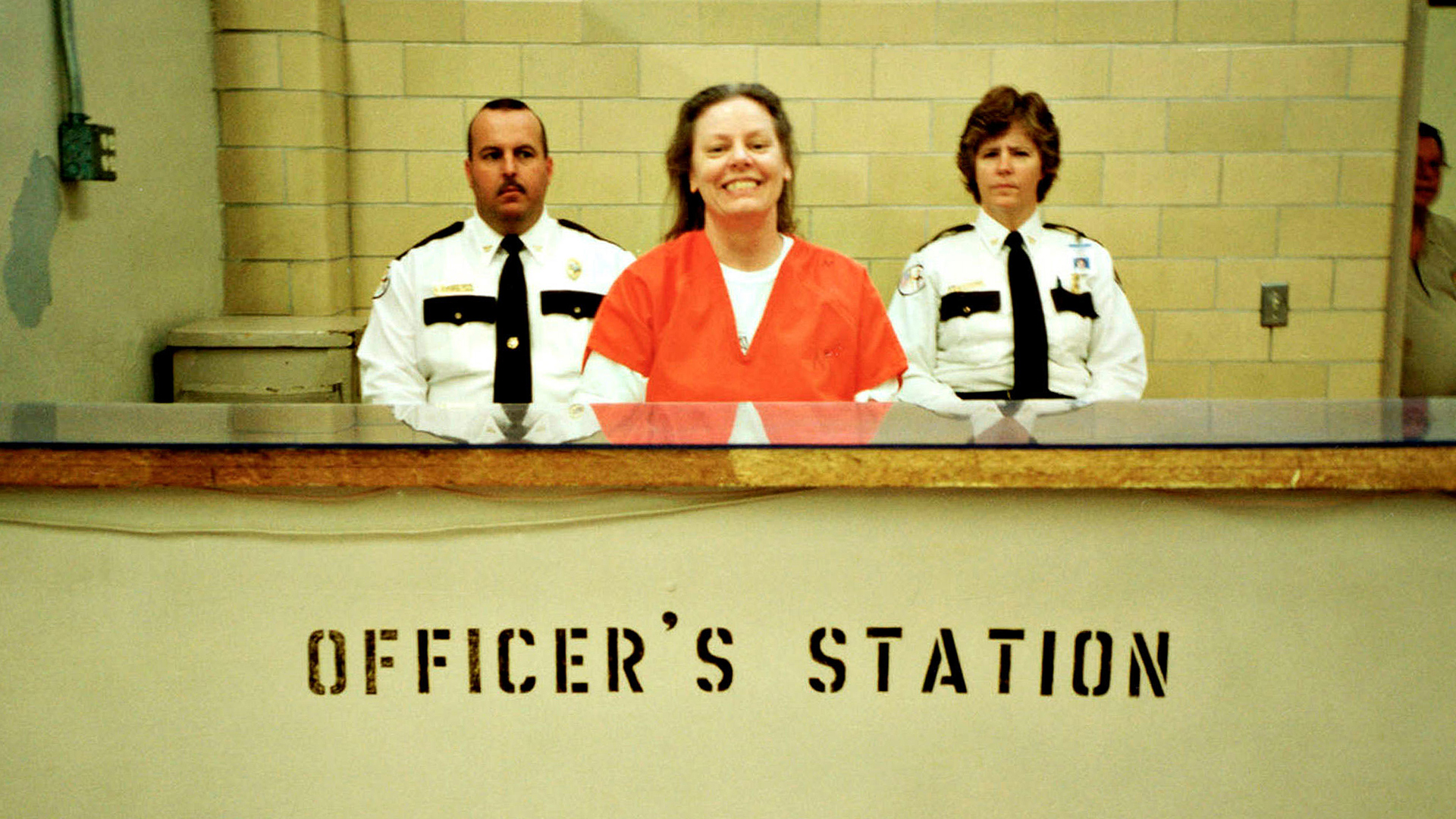 Aileen Wuornos smiling in an orange prison jumpsuit at the officer's station