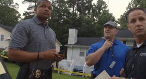 Three law enforcement officers stand by yellow police tape looking concerned