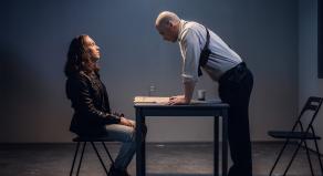 A policeman charges a man detained in handcuffs while inside an interrogation room.