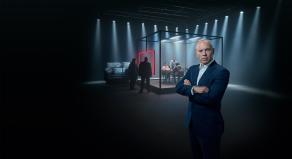 Rob Rinder standing with his arms crossed in front of a large, clear, cube