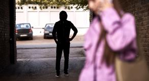 A hooded figure stands in an alleyway watching a woman walk past
