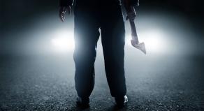 A man holds an axe in front of car headlights