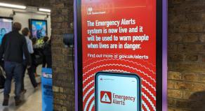 A photograph of a digital poster at London Waterloo train station, promoting the UK's Emergency Alerts system. 