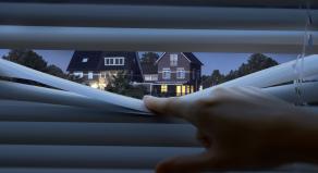 A photo showing a person peeking through some blinds, looking at a house