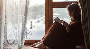 A woman reading a book sitting on a windowsill - a wintery scene is outside