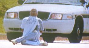 Screengrab of a local news report showing Brian Wells sitting cross-legged in front of a police car. His hands are cuffed behind his back and he has a bomb around his neck