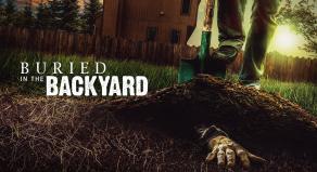 A hand appears the lawn in a backyard while a man stands with a shovel above it