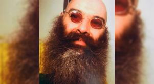 A close up image of Charles Bronson with a large beard and red-tinted sunglasses