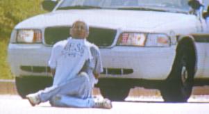 Screengrab of a local news report showing Brian Wells sitting cross-legged in front of a police car. His hands are cuffed behind his back and he has a bomb around his neck