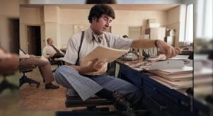 Colourised image of Dave Toschi looking through stacks of papers at his desk. He is wearing suspenders, a bow tie and tartan trousers