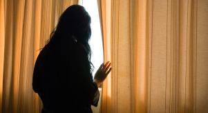 A woman cautiously looking out of her closed curtains