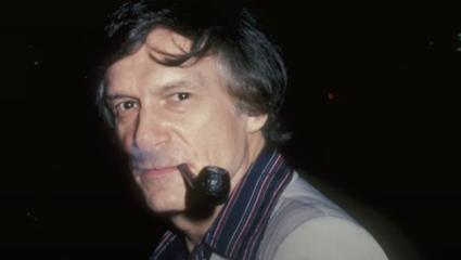 An old photo of Hugh Hefner with dark hair smoking a pipe