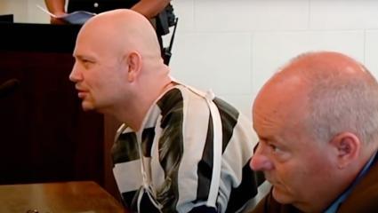 A bald prisoner wearing an arm sling leans forward in court