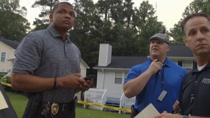Three law enforcement officers stand by yellow police tape looking concerned