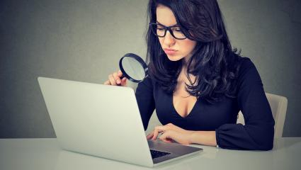 A woman in glasses examines her laptop screen with a magnifying glass