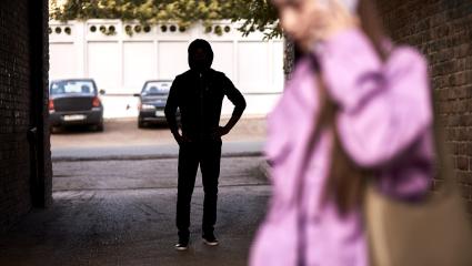 A hooded figure stands in an alleyway watching a woman walk past