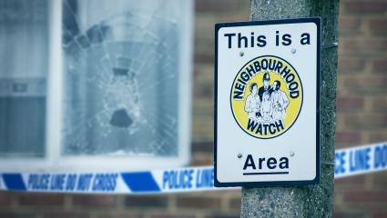 A stock image of a smashed window in front of a Neighbourhood Watch sign