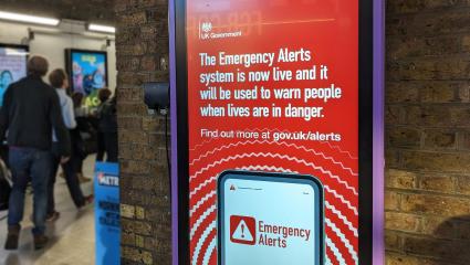 A photograph of a digital poster at London Waterloo train station, promoting the UK's Emergency Alerts system. 