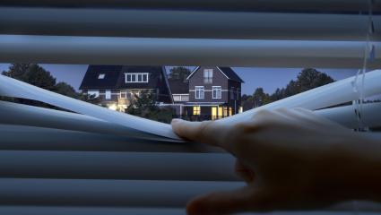 A photo showing a person peeking through some blinds, looking at a house