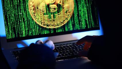A hacker working on a laptop as their screen shows binary code and the Bitcoin logo