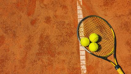 Three tennis balls and a tennis racquet lie on a clay tennis court