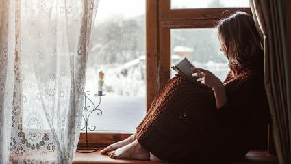 A woman reading a book sitting on a windowsill - a wintery scene is outside