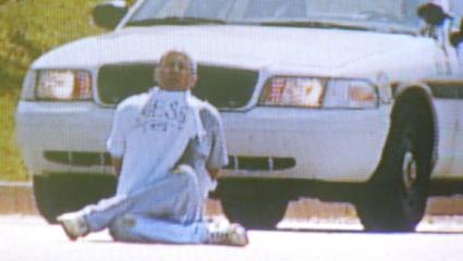 Screengrab of a local news report showing Brian Wells sitting cross-legged in front of a police car. His hands are cuffed behind his back and he has a bomb around his neck
