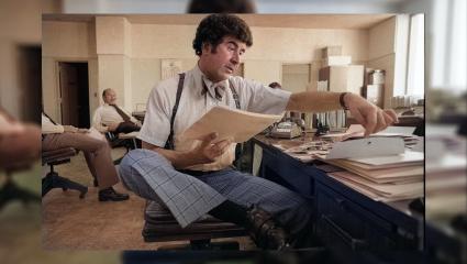 Colourised image of Dave Toschi looking through stacks of papers at his desk. He is wearing suspenders, a bow tie and tartan trousers