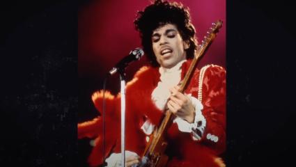 Prince wearing a red suit on stage and playing a guitar