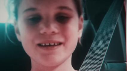 A young Holly Newton smiles slightly while sitting in a car, wearing a seatbelt. The photo appears to be taken from a video or screen, with a soft, bluish tint.