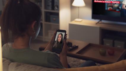 A woman looks at an image of True Crime Caitlyn on her phone while 'Laura Whitmore on Britain's Killer Teens' plays on her television