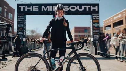 A photography of Anna Moriah Wilson posing with her bicycle at a race. In the background of the photo is a starting gantry, with spectators also visible either side of barriers