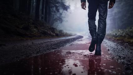 A man's legs as he walks along a road in the rain. A puddle on the road appears red