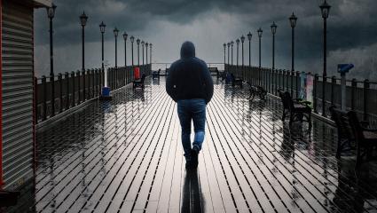 A man with his hood up walking away from the camera along a rainy pier