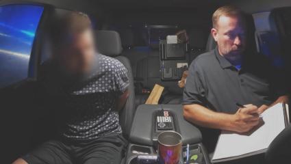 A predator with their face blurred speaks to a police officer in the back of a car