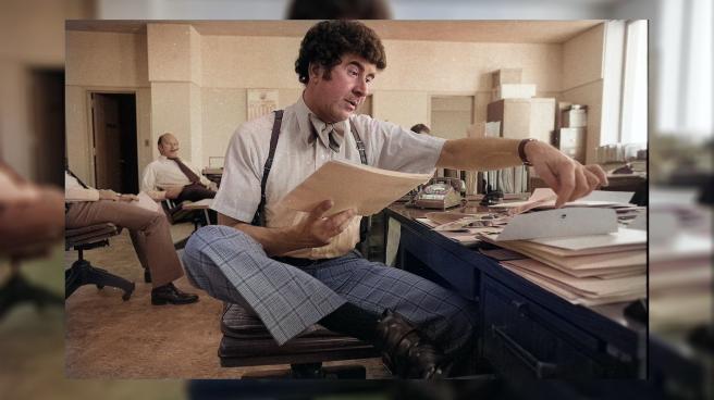 Colourised image of Dave Toschi looking through stacks of papers at his desk. He is wearing suspenders, a bow tie and tartan trousers