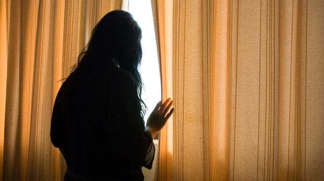 A woman cautiously looking out of her closed curtains