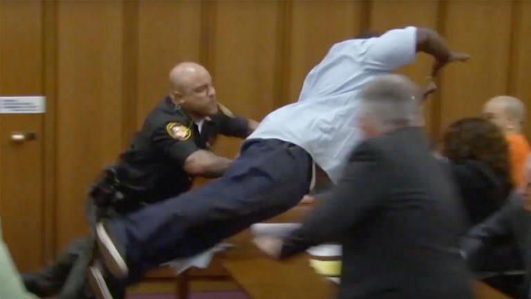 A man jumps over a table in court while a policeman tries to stop him