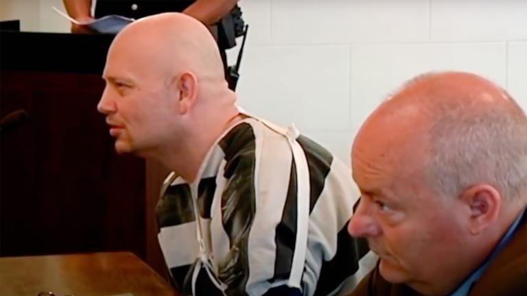A bald prisoner wearing an arm sling leans forward in court