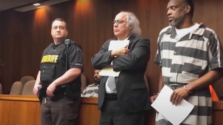 Ralph Butler in court wearing his black and white jumpsuit next to his lawyer and a police guard
