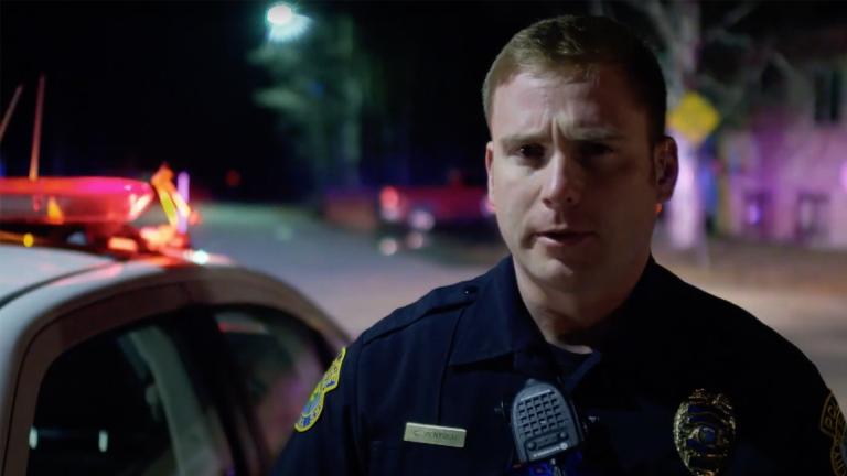 A police officer talking to the camera in front of a police car at a traffic stop