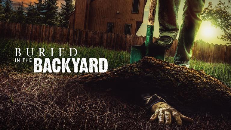 A hand appears the lawn in a backyard while a man stands with a shovel above it