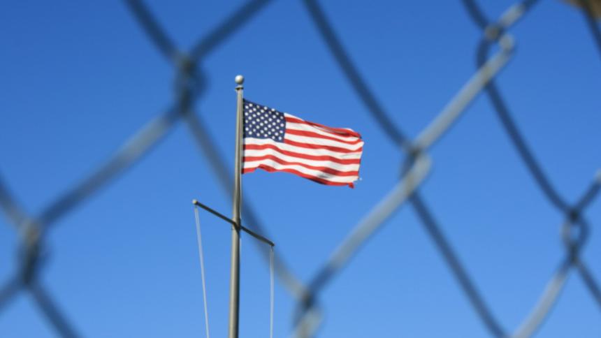 A photograph of the American flag behind a chain link fence