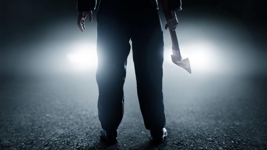 A man holds an axe in front of car headlights