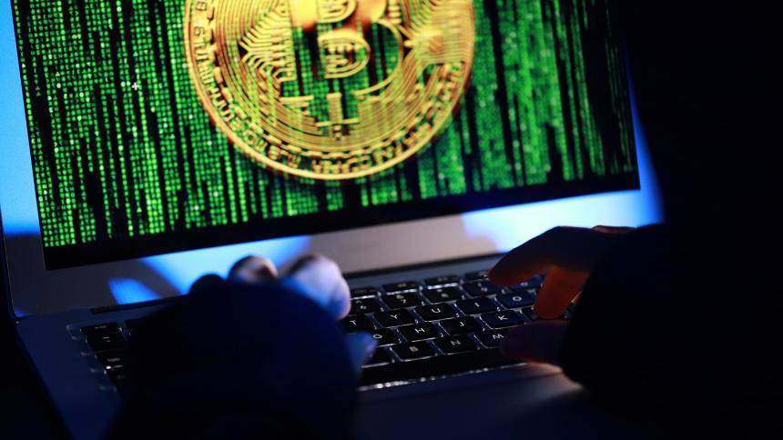 A hacker working on a laptop as their screen shows binary code and the Bitcoin logo